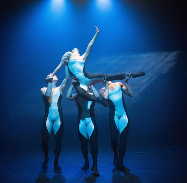 Three dancers lift a female dancer. Her back leg extends while her front leg tucks underneath her pelvis.